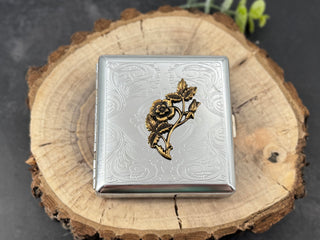 A silver-toned metal cigarette case with an embossed floral design and a raised gold-toned rose motif on the front, displayed on a round wood slice against a dark gray background.