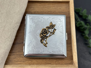 A silver-toned metal cigarette case with an embossed floral scroll pattern and a raised gold-toned flower motif on the front, displayed on a wooden tray with a beige fabric napkin and green leaves in the background.