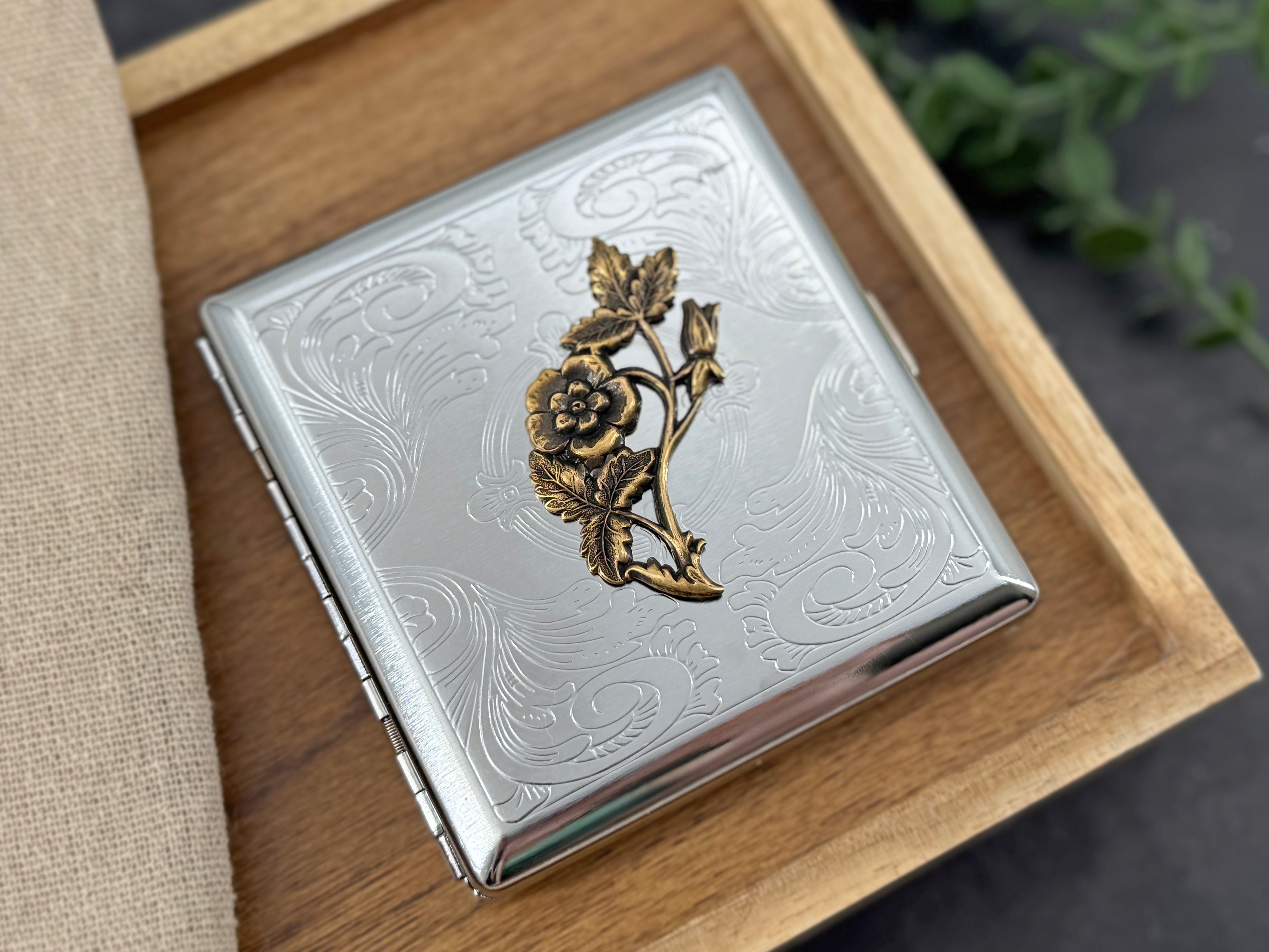A silver-toned metal cigarette case with an embossed wave pattern and a gold-toned rose branch decoration on the front, resting on a wooden tray.