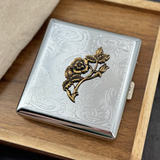 A square silver-toned metal case with embossed floral scrollwork and a raised antique-gold flower branch motif on the front, resting on a wooden tray with a beige fabric corner visible.