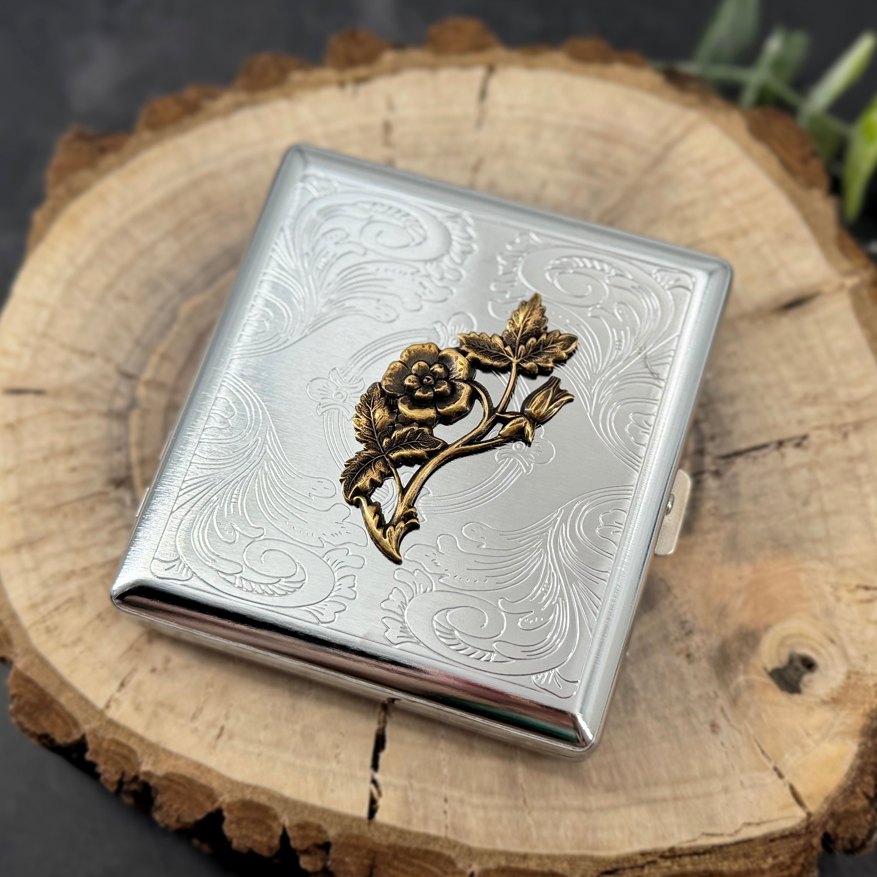 A silver-toned metal cigarette case with an engraved floral design and a raised gold-toned rose motif on the front, displayed on a rustic wood slice background.