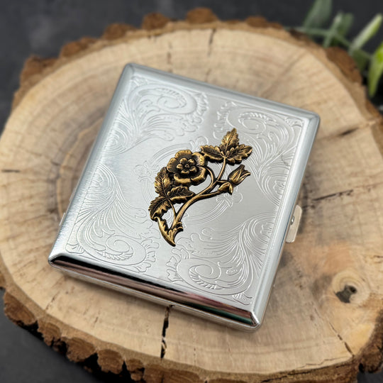 A silver-toned metal cigarette case with an engraved floral design and a raised gold-toned rose motif on the front, displayed on a rustic wood slice background.