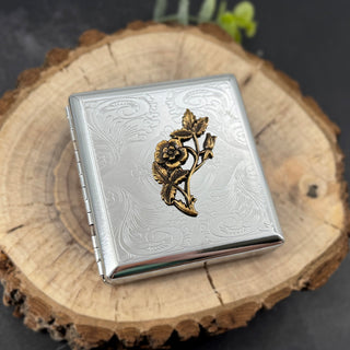 A square silver-toned metal case with engraved floral scrollwork and a raised gold-toned rose motif on the front, displayed on a round wood slice against a dark background.