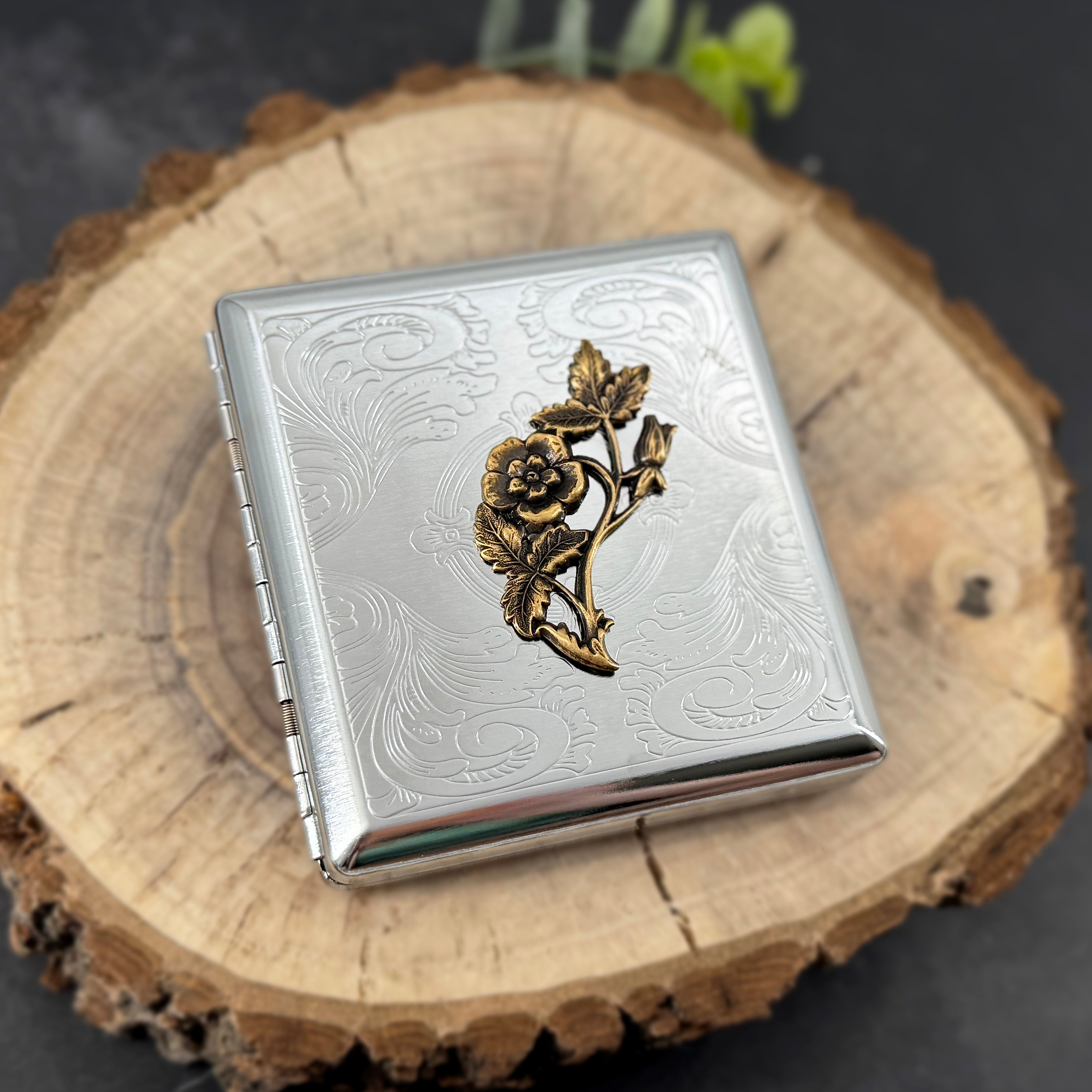A square silver-toned metal case with engraved floral scrollwork and a raised gold-toned rose motif on the front, displayed on a round wood slice against a dark background.