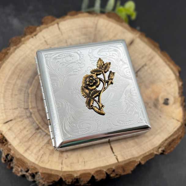 A square silver-toned metal case with engraved floral scrollwork and a raised gold-toned rose motif on the front, displayed on a round wood slice against a dark background.