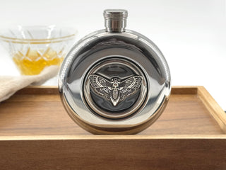 A polished silver metal flask with a winged insect emblem on the front, displayed on a wooden tray with a glass of amber liquid blurred in the background.