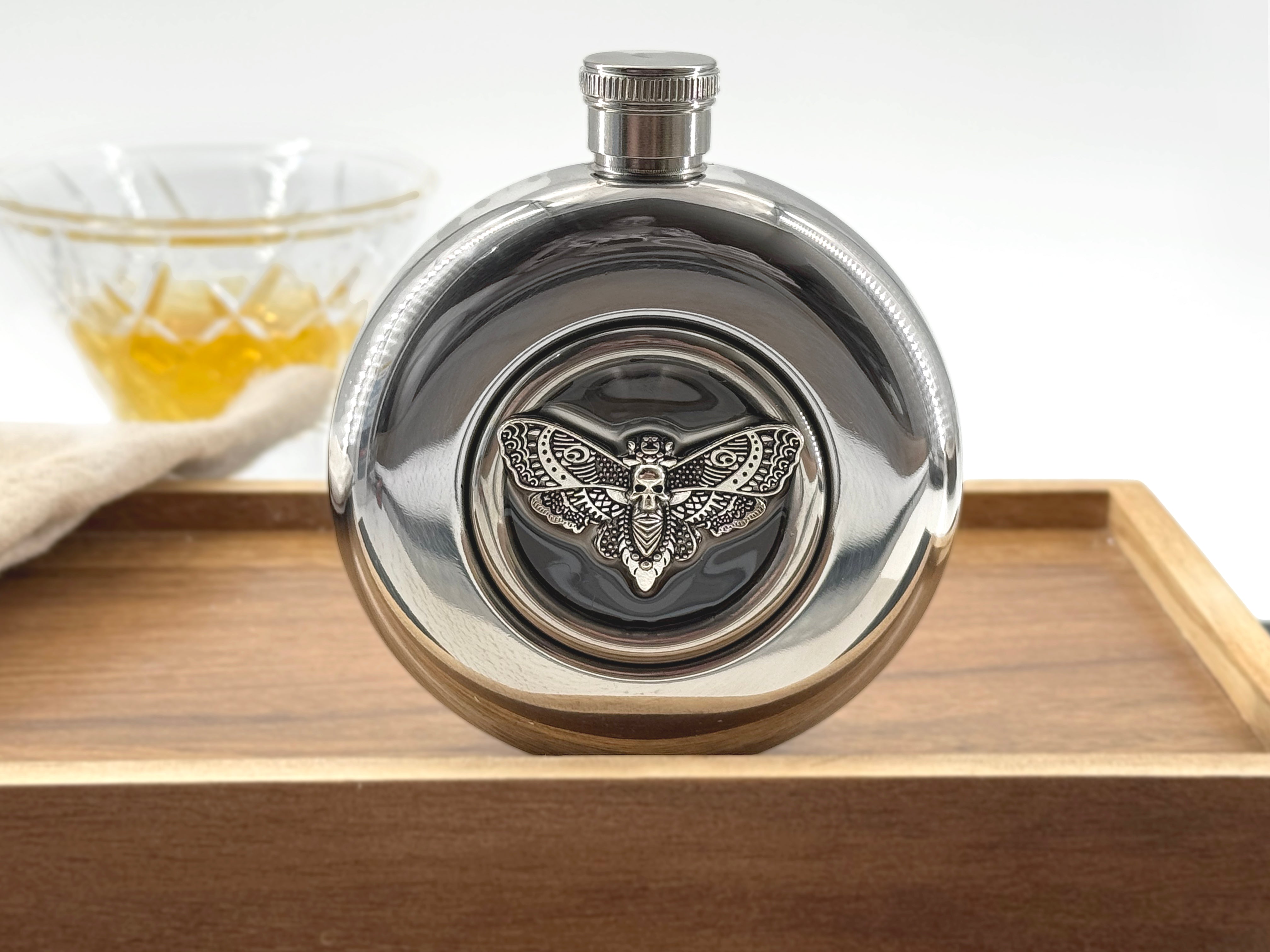 A polished silver metal flask with a winged insect emblem on the front, displayed on a wooden tray with a glass of amber liquid blurred in the background.