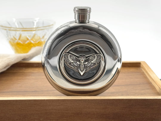 A polished silver metal flask with a winged insect emblem on the front, displayed on a wooden tray with a glass of amber liquid blurred in the background.