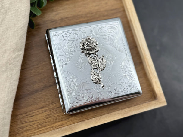 Silver cigarette case featuring a raised rose on an ornate stamped scrollwork background.