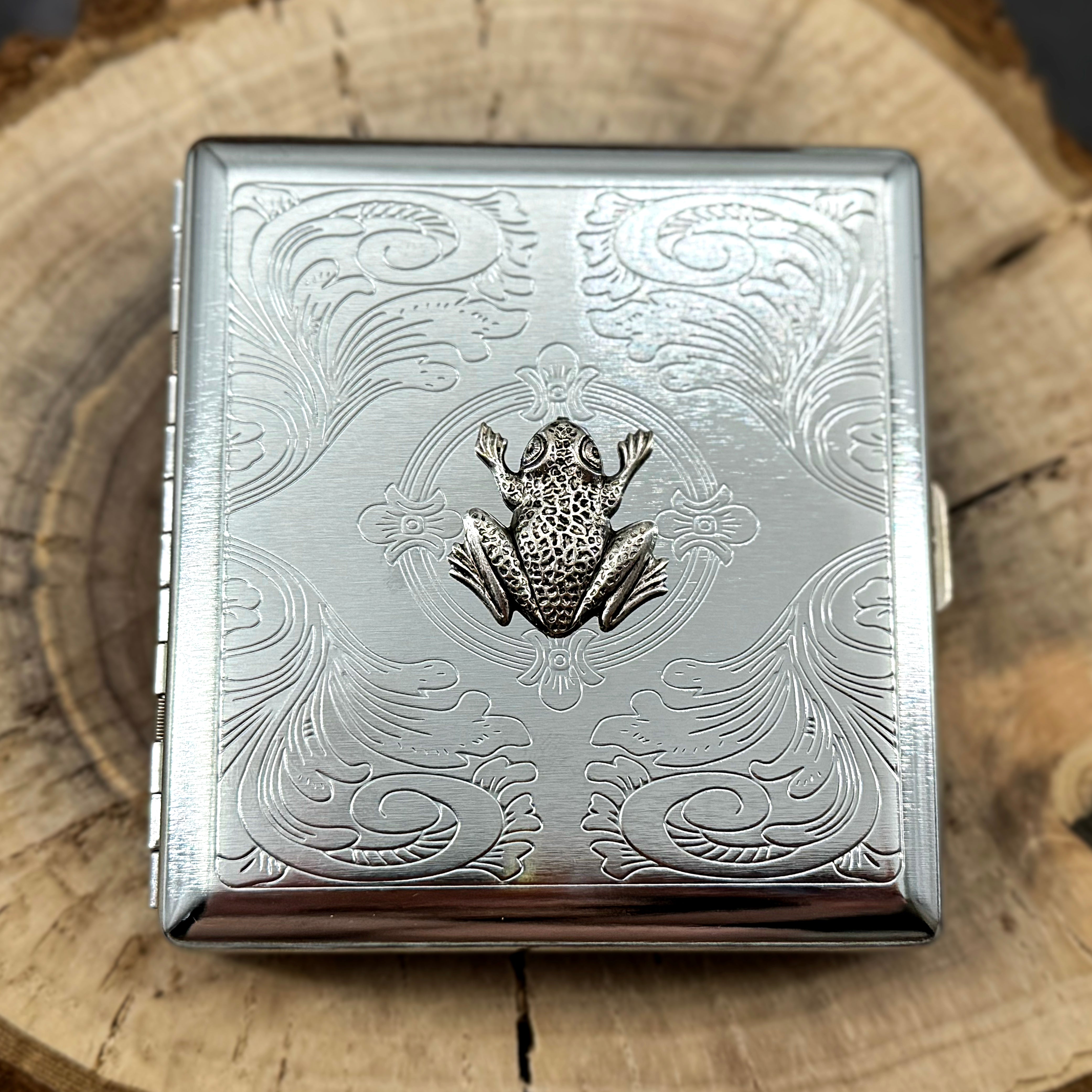 Silver cigarette case featuring a raised frog on a stamped metal scrollwork background.