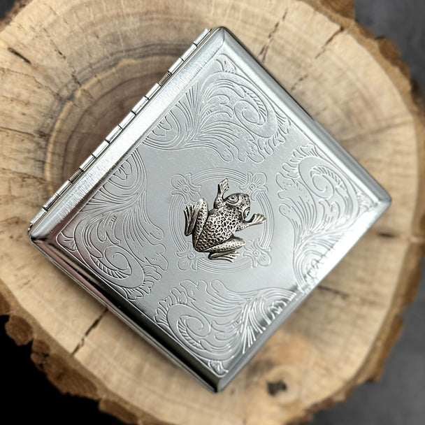 Metal cigarette case with a raised frog and ornate stamped metal scrollwork.
