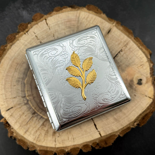 Silver cigarette case with raised gold leaf design on a stamped metal scrollwork background.
