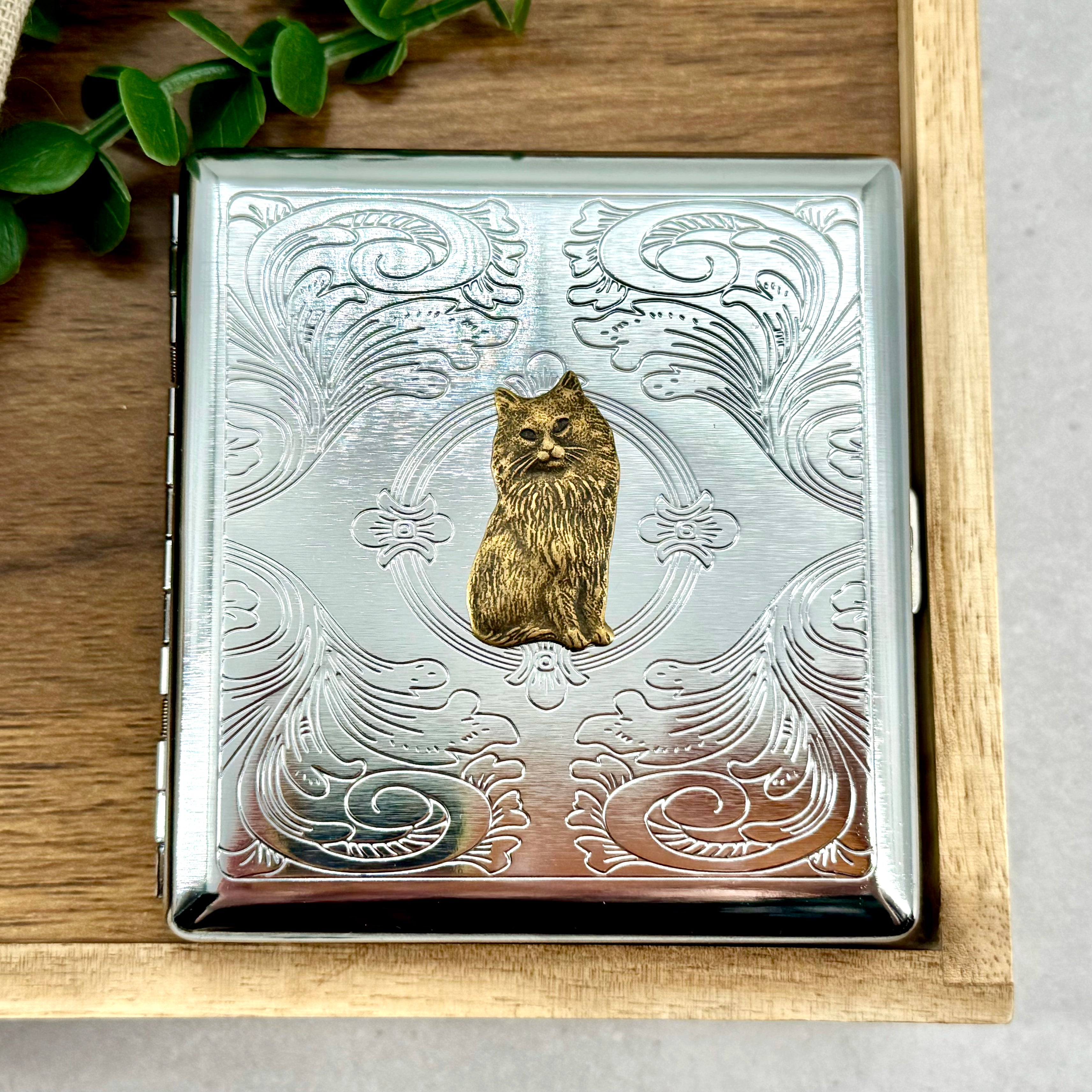 Silver stamped metal cigarette case with raised Persian cat charm on ornate background