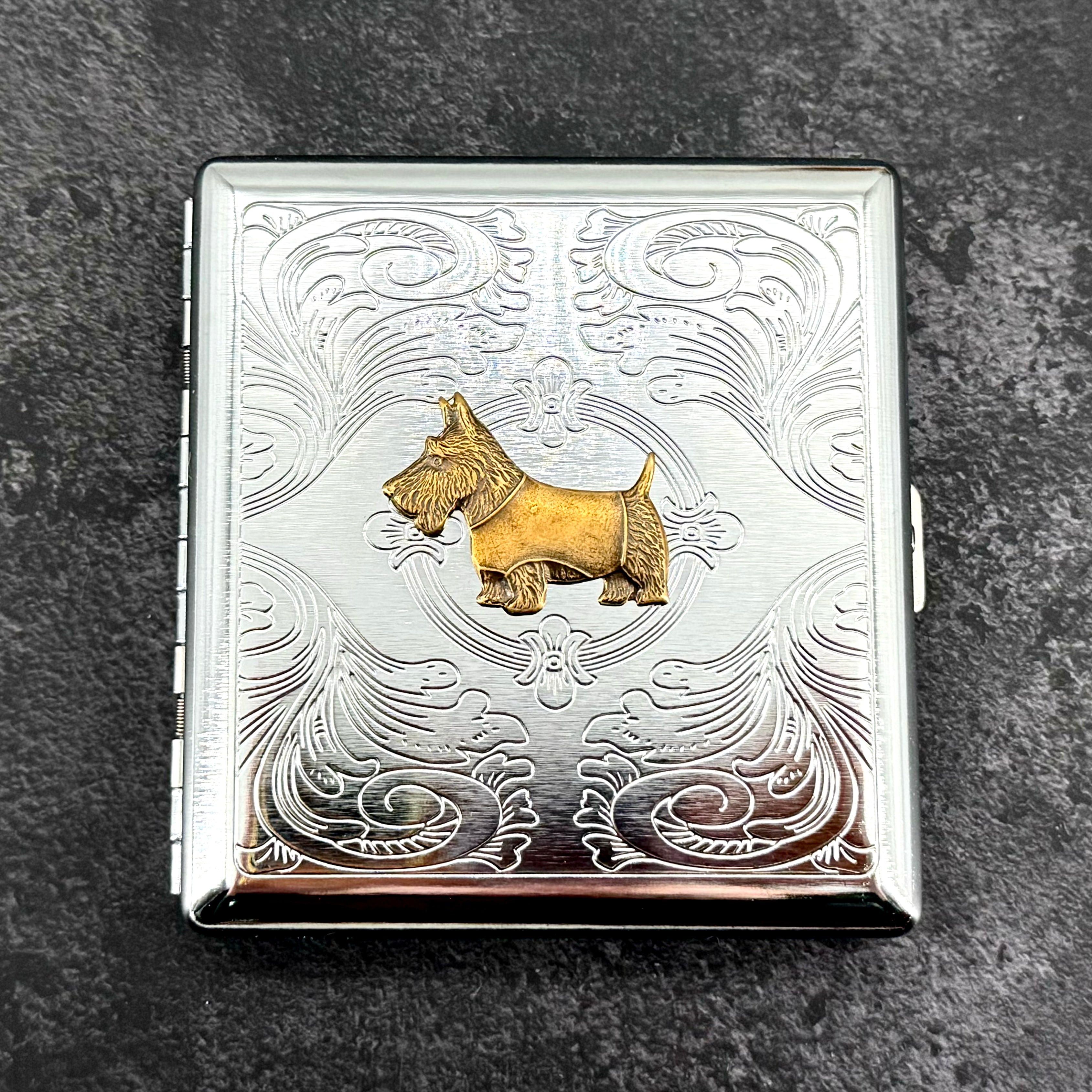 Angled view of Scottie dog metal cigarette case with ornate stamped scrollwork background
