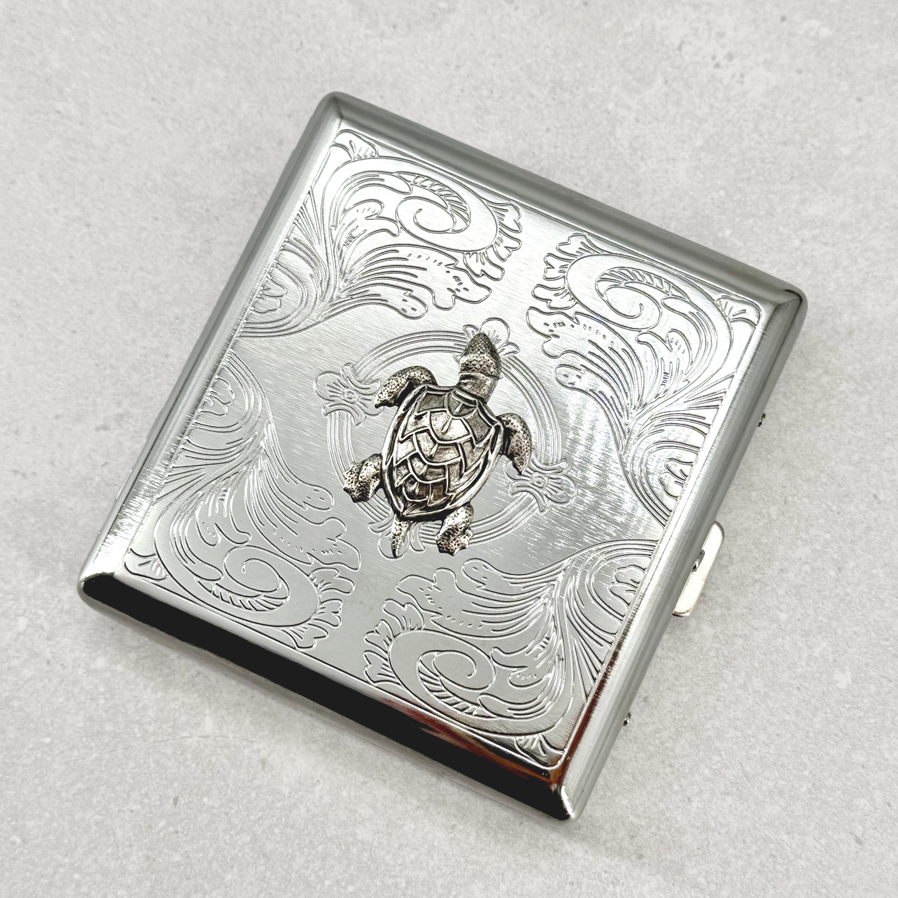 Angled view of sea turtle metal cigarette case with ornate stamped scrollwork design