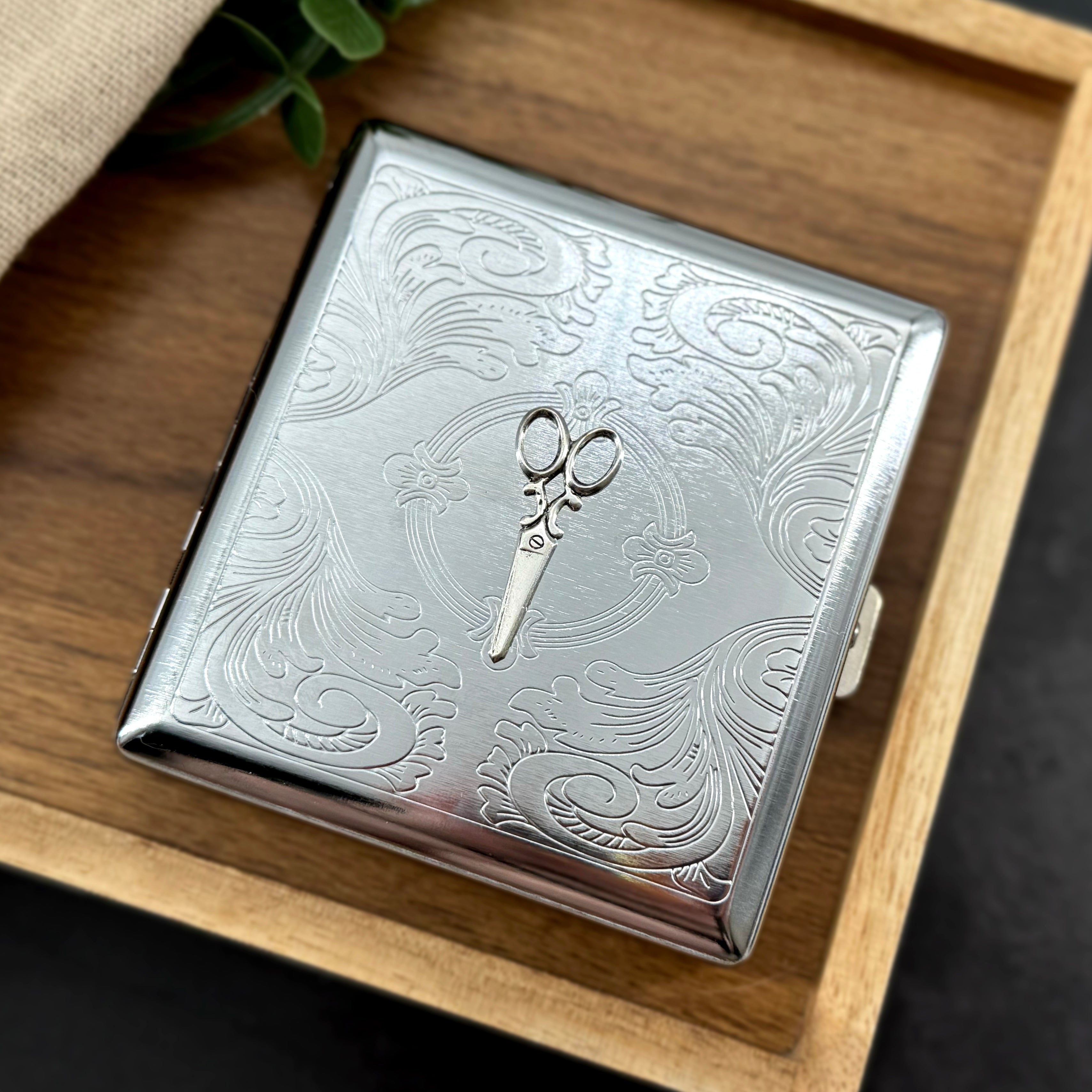 Scissors metal cigarette case featuring raised silver shears on an ornate stamped scrollwork background
