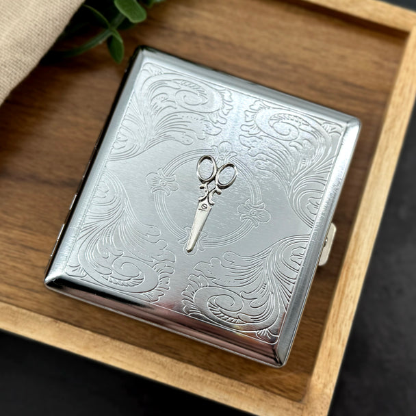 Scissors metal cigarette case featuring raised silver shears on an ornate stamped scrollwork background
