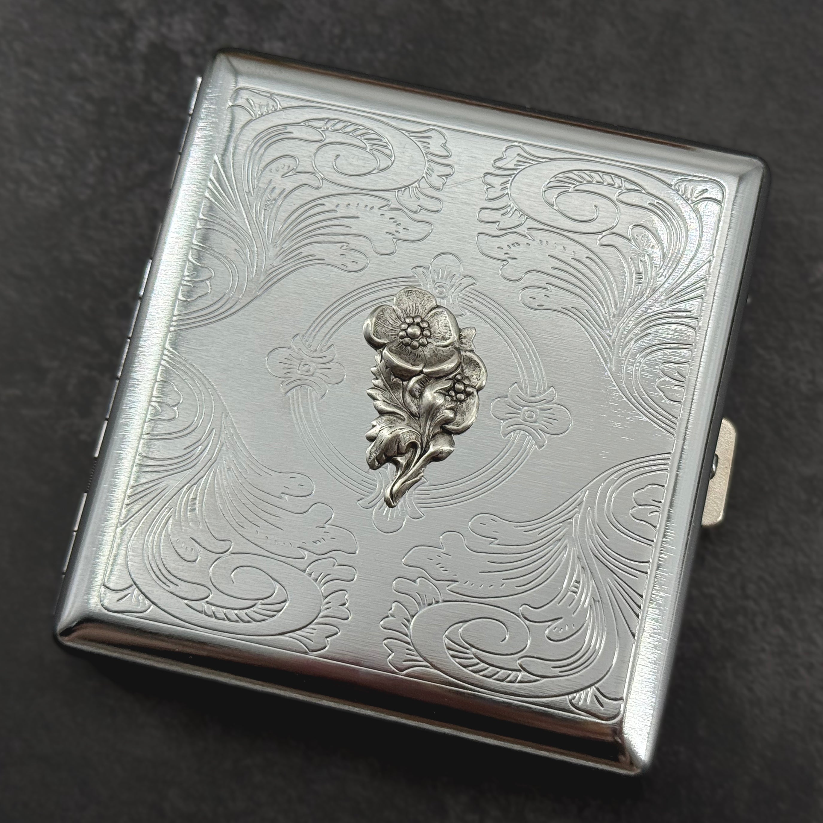 Closed silver metal cigarette case featuring an ornate geometric stamped border and textured center panel
