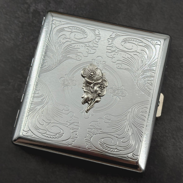 Closed silver metal cigarette case featuring an ornate geometric stamped border and textured center panel