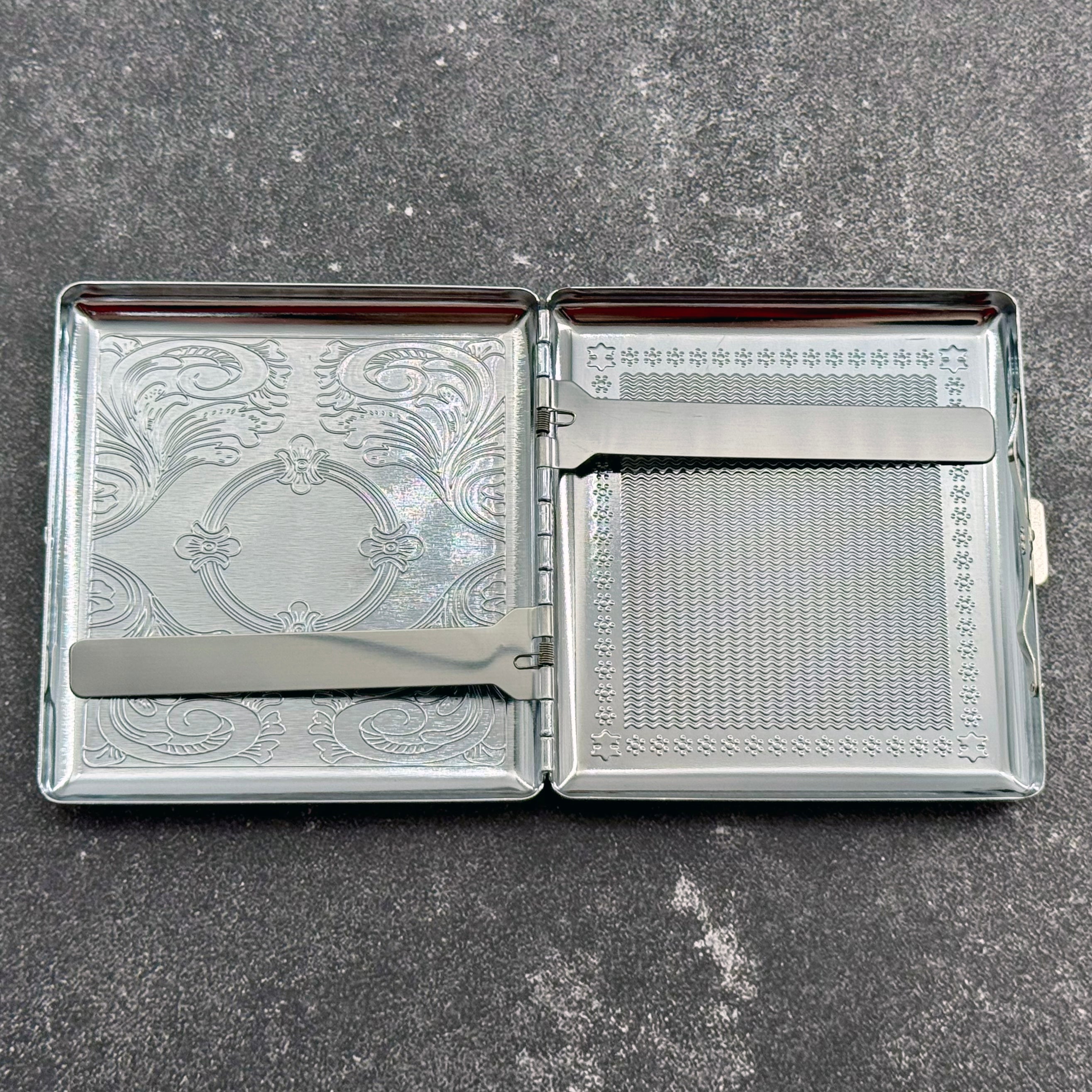 Open silver cigarette case showing the smooth interior and two retaining arms that help keep cigarettes, cards, cash, or other small items in place.